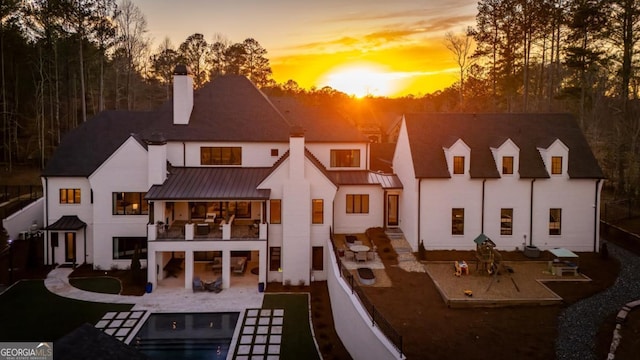 back of house at dusk featuring a patio, a chimney, a standing seam roof, metal roof, and a balcony