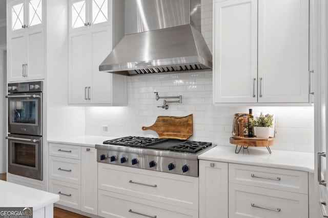 kitchen featuring glass insert cabinets, stainless steel appliances, light countertops, wall chimney range hood, and white cabinetry