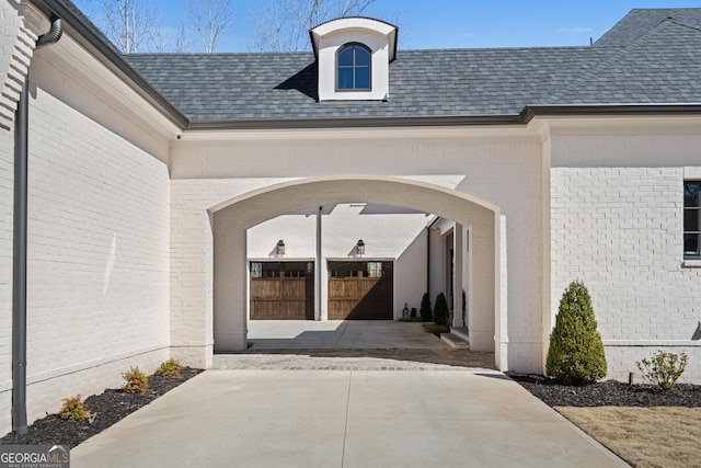 entrance to property featuring concrete driveway, brick siding, and roof with shingles