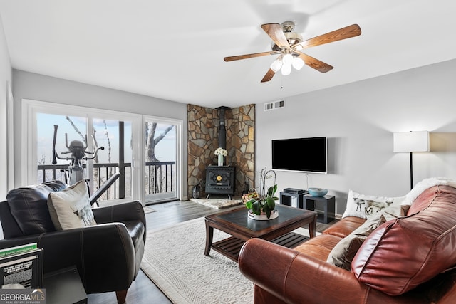 living room with a wood stove, wood finished floors, visible vents, and a ceiling fan