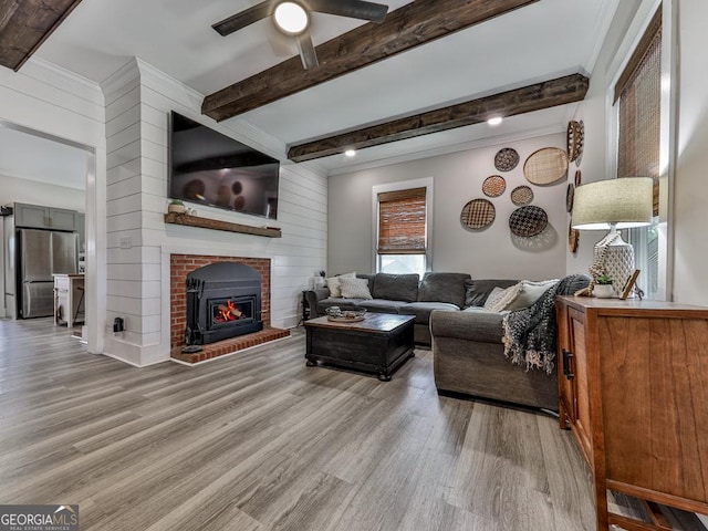 living area featuring ceiling fan, beamed ceiling, light wood-type flooring, and a brick fireplace