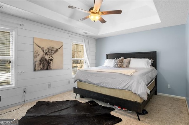 bedroom featuring ceiling fan, wooden walls, light colored carpet, baseboards, and a tray ceiling