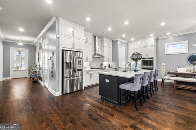 kitchen featuring light countertops, appliances with stainless steel finishes, a kitchen island with sink, white cabinetry, and wall chimney exhaust hood