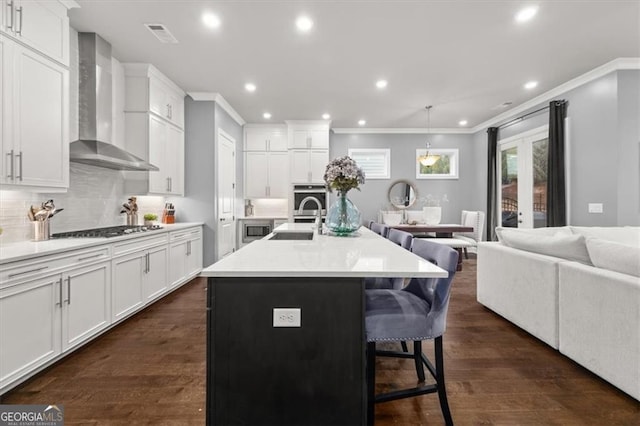 kitchen featuring wall chimney range hood, a kitchen island with sink, light countertops, and open floor plan