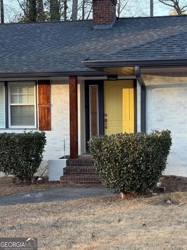 doorway to property with roof with shingles, a chimney, and brick siding