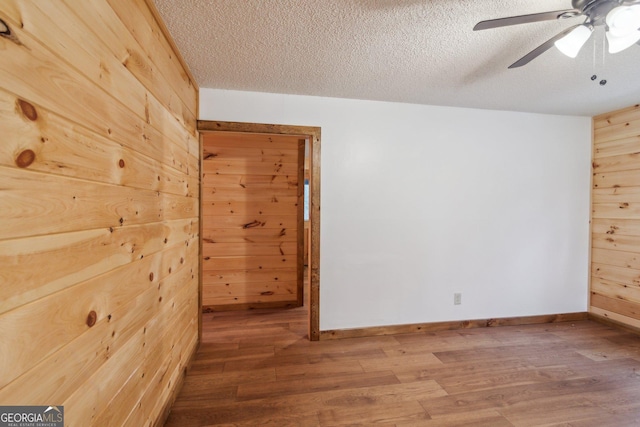 empty room with a textured ceiling, wood walls, wood finished floors, a ceiling fan, and baseboards