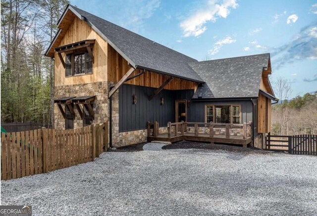 rustic home with a deck, stone siding, a shingled roof, and fence