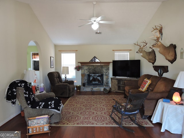 living area with arched walkways, dark wood-type flooring, a ceiling fan, a stone fireplace, and high vaulted ceiling