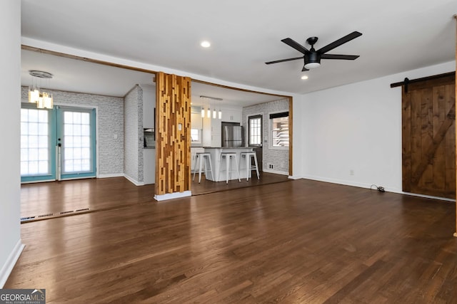 unfurnished living room with ceiling fan with notable chandelier, a barn door, dark wood-type flooring, and baseboards