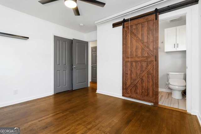 unfurnished bedroom with a barn door, connected bathroom, dark wood-type flooring, a ceiling fan, and baseboards