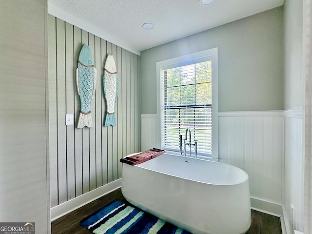 full bath featuring a textured ceiling, wood finished floors, a freestanding bath, and wainscoting