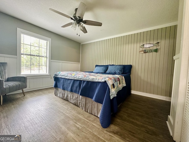 bedroom with a textured ceiling, ceiling fan, a wainscoted wall, and dark wood-style floors