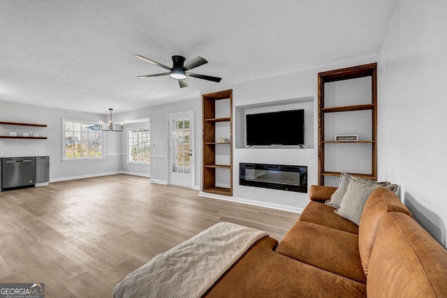 living area featuring baseboards, a glass covered fireplace, wood finished floors, built in shelves, and ceiling fan with notable chandelier