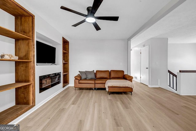 living area featuring light wood finished floors, built in features, baseboards, a glass covered fireplace, and ceiling fan