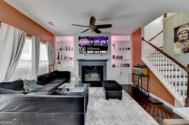 living room with dark wood-type flooring, visible vents, a ceiling fan, stairway, and a glass covered fireplace