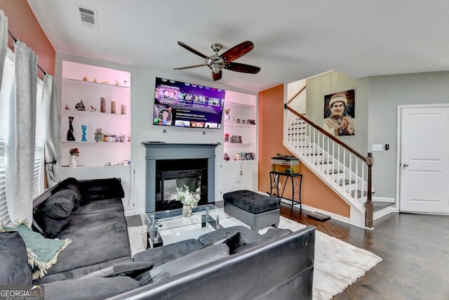 living room with ceiling fan, visible vents, stairway, dark wood-style floors, and a glass covered fireplace