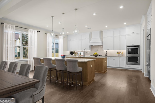 kitchen featuring a kitchen island with sink, premium range hood, white cabinets, light countertops, and pendant lighting