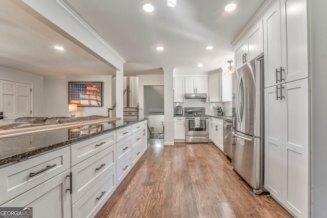 kitchen with under cabinet range hood, white cabinetry, appliances with stainless steel finishes, dark stone counters, and crown molding
