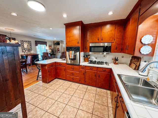 kitchen with stainless steel appliances, a peninsula, a sink, tile counters, and tasteful backsplash