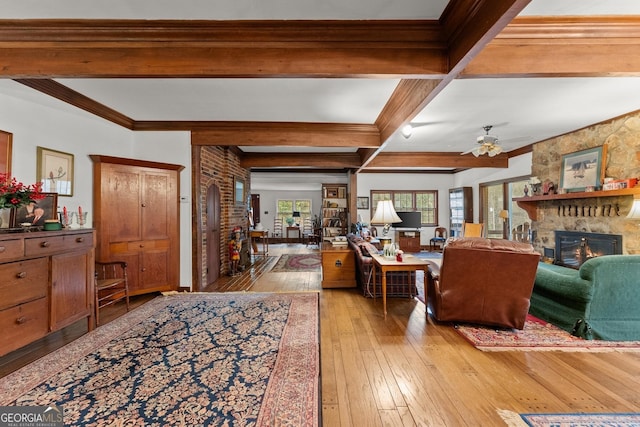 living room with beam ceiling, light wood finished floors, ornamental molding, ceiling fan, and a stone fireplace