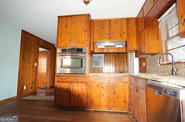 kitchen with under cabinet range hood, dark wood-type flooring, a sink, appliances with stainless steel finishes, and brown cabinetry