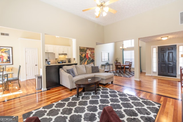 living room featuring ceiling fan with notable chandelier, a high ceiling, visible vents, baseboards, and light wood finished floors