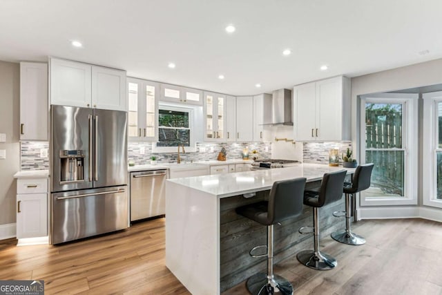 kitchen featuring a breakfast bar area, stainless steel appliances, light countertops, white cabinetry, and wall chimney range hood