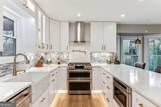 kitchen with stainless steel appliances, a sink, white cabinets, wall chimney range hood, and light wood-type flooring