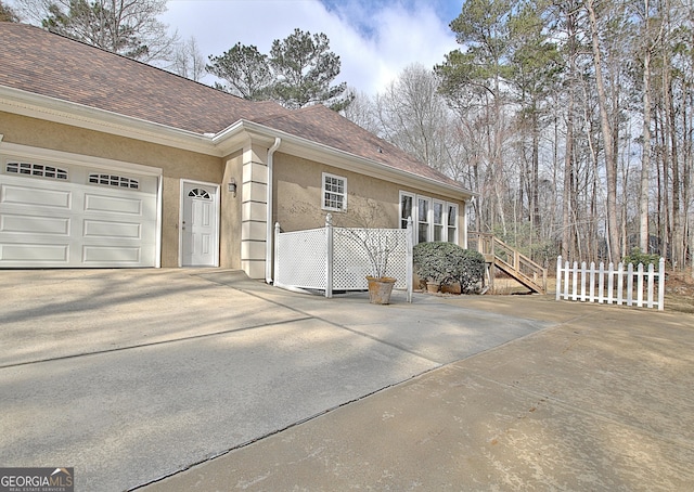 view of home's exterior with driveway, roof with shingles, an attached garage, fence, and stucco siding