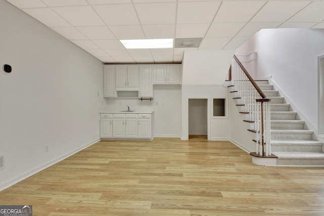 unfurnished living room with a paneled ceiling, light wood finished floors, baseboards, and stairway