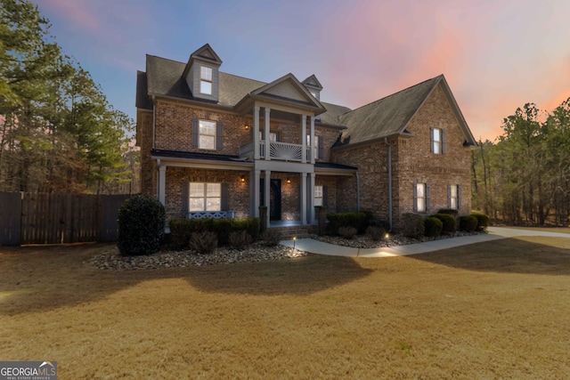 traditional-style home featuring a balcony, fence, driveway, a front lawn, and brick siding