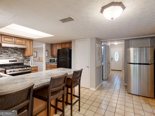 kitchen featuring tasteful backsplash, visible vents, stainless steel appliances, under cabinet range hood, and a kitchen bar