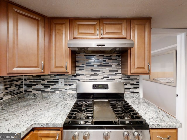 kitchen with brown cabinets, decorative backsplash, stainless steel gas stove, light stone countertops, and under cabinet range hood