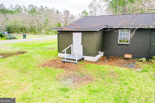 rear view of property featuring entry steps, a shingled roof, concrete block siding, and a lawn