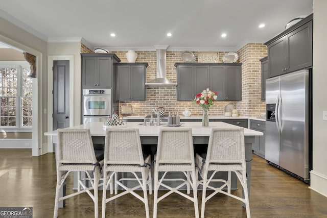 kitchen with a breakfast bar area, a kitchen island with sink, stainless steel appliances, light countertops, and wall chimney range hood