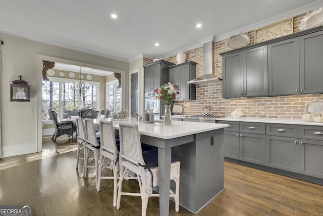 kitchen featuring a breakfast bar, a kitchen island with sink, light countertops, gray cabinetry, and wall chimney range hood