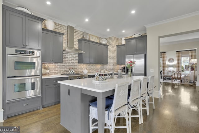 kitchen with a breakfast bar area, gray cabinets, light countertops, appliances with stainless steel finishes, and wall chimney exhaust hood