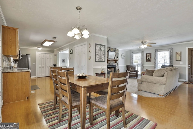 dining room featuring light wood finished floors, a stone fireplace, ceiling fan with notable chandelier, and ornamental molding