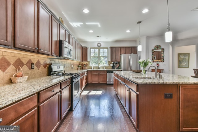 kitchen featuring a center island with sink, appliances with stainless steel finishes, light stone counters, hanging light fixtures, and a sink