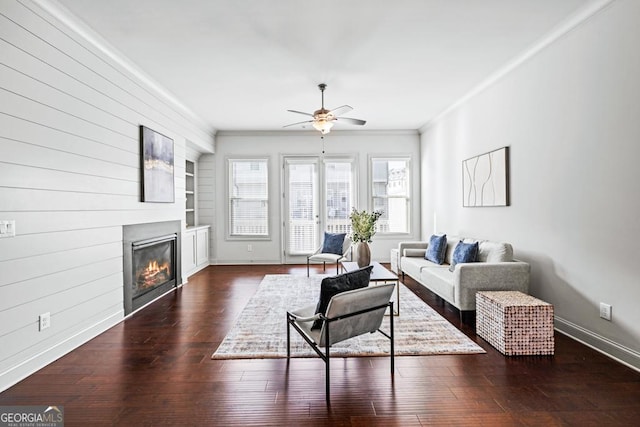 living area featuring built in shelves, dark wood finished floors, ornamental molding, a glass covered fireplace, and baseboards