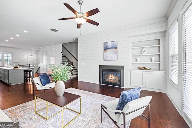 living room featuring built in shelves, a glass covered fireplace, visible vents, and dark wood finished floors