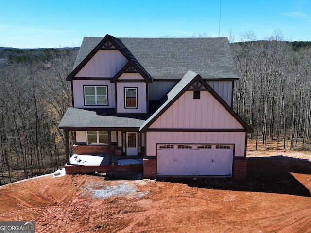 view of front facade with brick siding, a shingled roof, covered porch, an attached garage, and a forest view