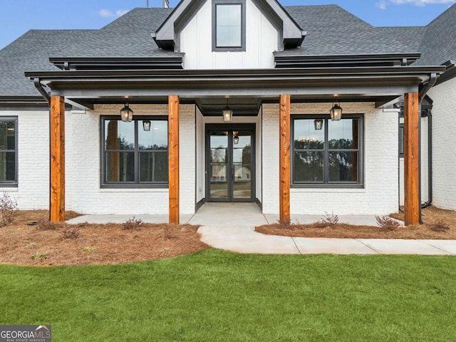 doorway to property featuring a yard, brick siding, a shingled roof, and french doors