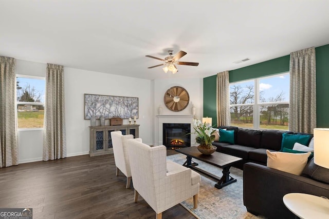 living room featuring visible vents, dark wood-type flooring, a glass covered fireplace, a ceiling fan, and baseboards