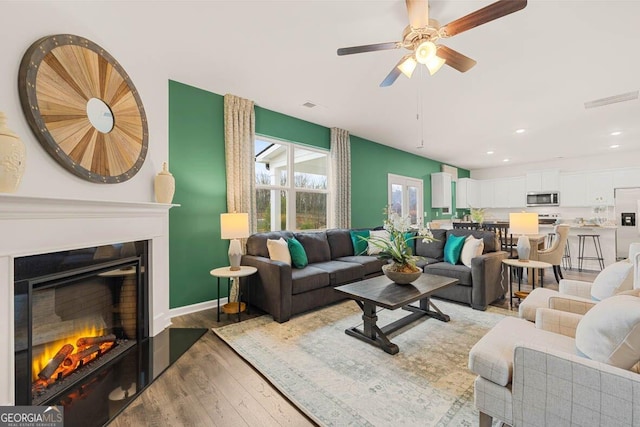 living area featuring visible vents, baseboards, a glass covered fireplace, ceiling fan, and wood finished floors