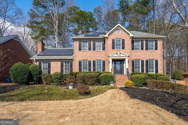 view of front facade with brick siding, a chimney, a front lawn, and roof mounted solar panels