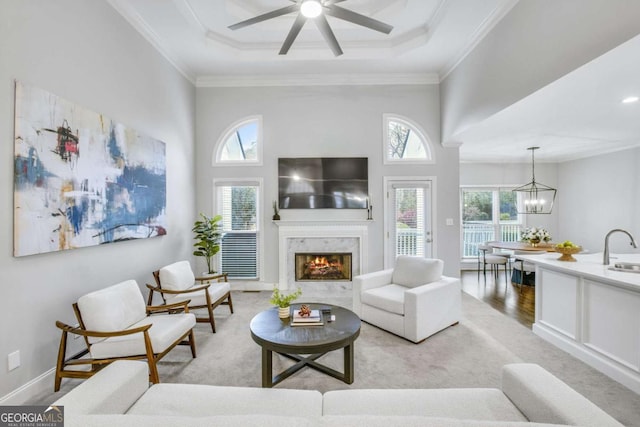 living area featuring baseboards, a raised ceiling, ornamental molding, a fireplace, and ceiling fan with notable chandelier