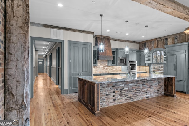 kitchen with light stone counters, built in appliances, custom exhaust hood, a large island with sink, and a sink