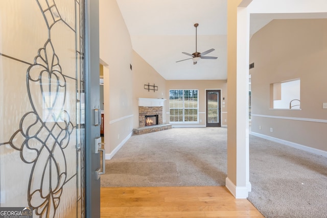 entryway featuring a ceiling fan, light carpet, a stone fireplace, high vaulted ceiling, and light wood-type flooring