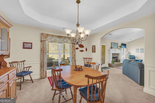 dining room with light carpet, arched walkways, a raised ceiling, a stone fireplace, and a chandelier